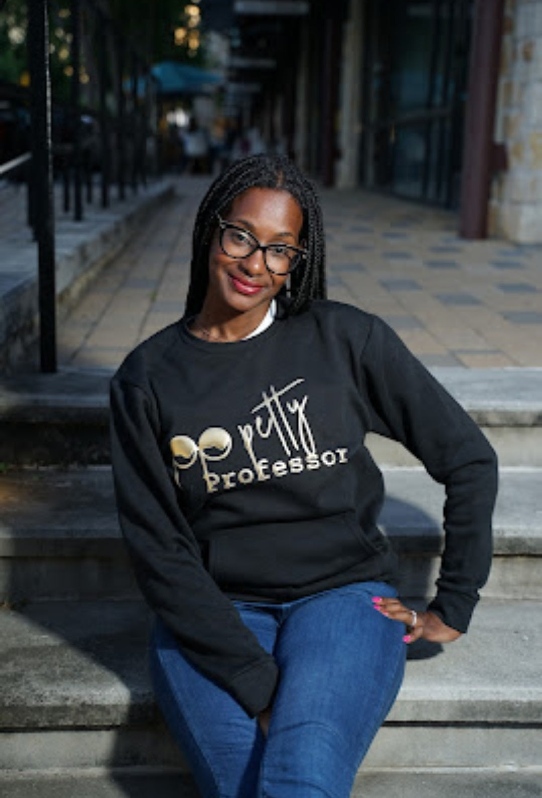 Woman wearing a black sweatshirt with 'pretty professor' text sitting on steps outdoors.